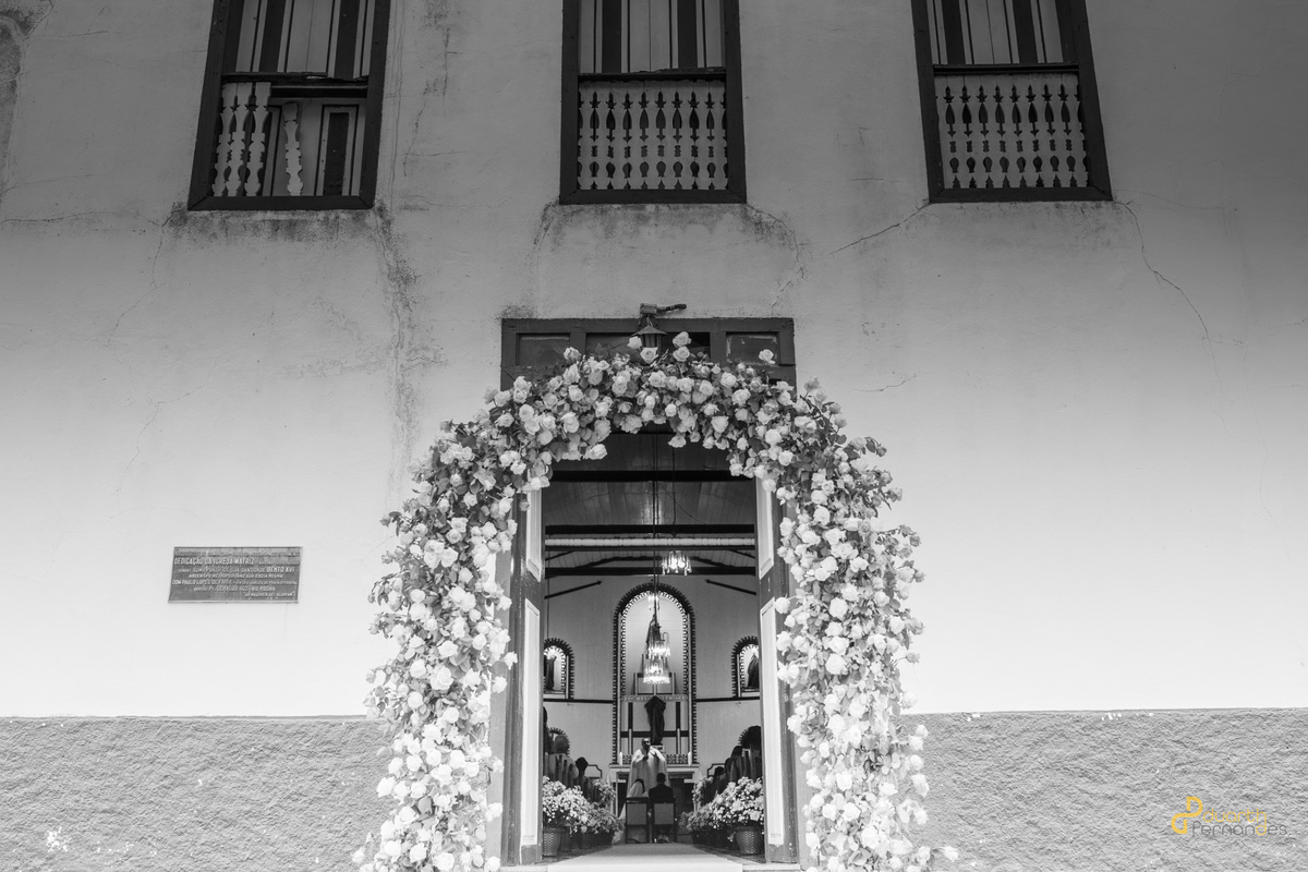 achada de igreja histórica em preto e branco com um grande arco decorativo de flores na porta de entrada durante o casamento de Gardênia e Arthur. Fotografia de casamento em Aricanduva, MG.