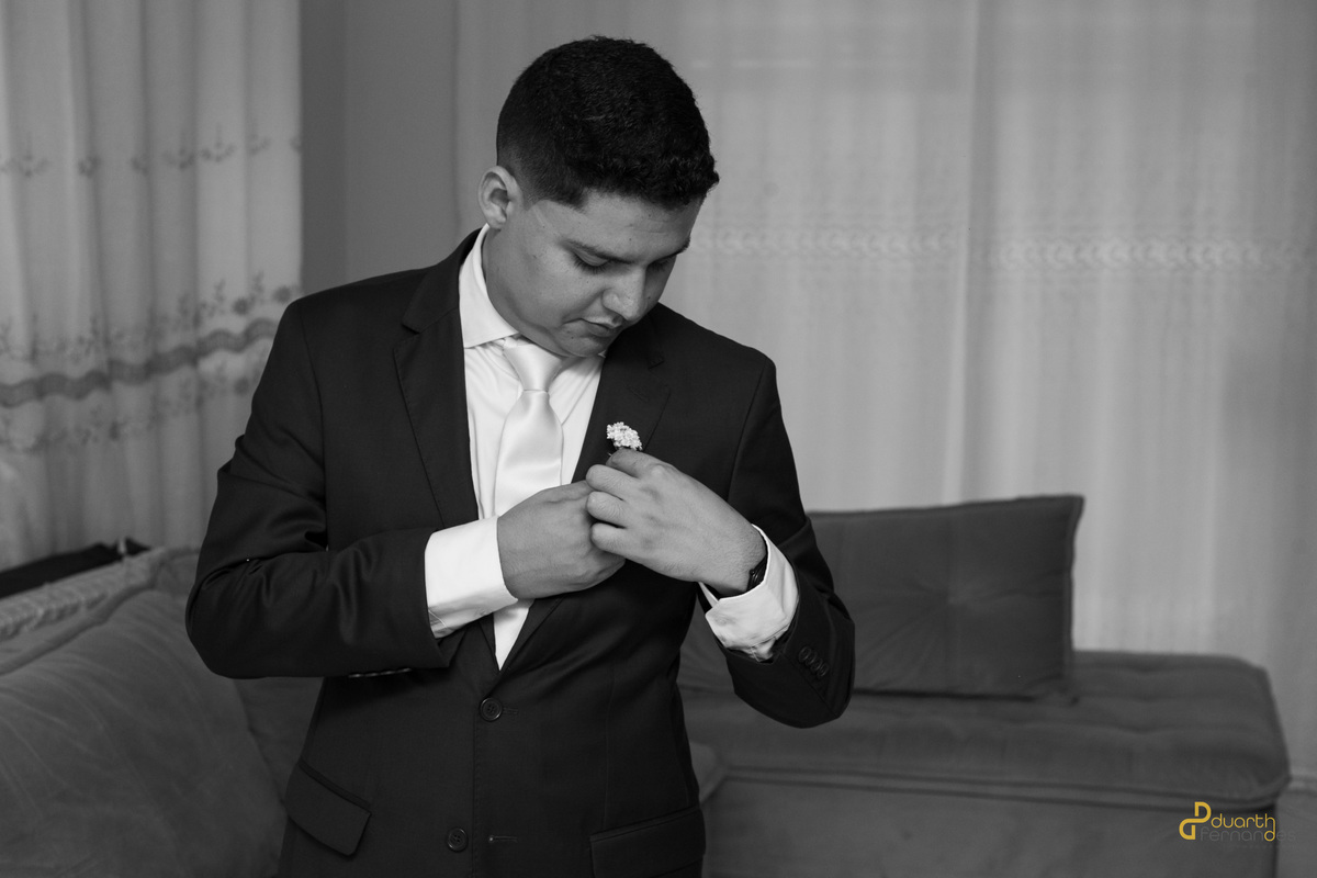 Foto em preto e branco do noivo Arthur arrumando a flor de lapela em seu terno preto antes da cerimônia. Ensaio making of de casamento em Aricanduva.