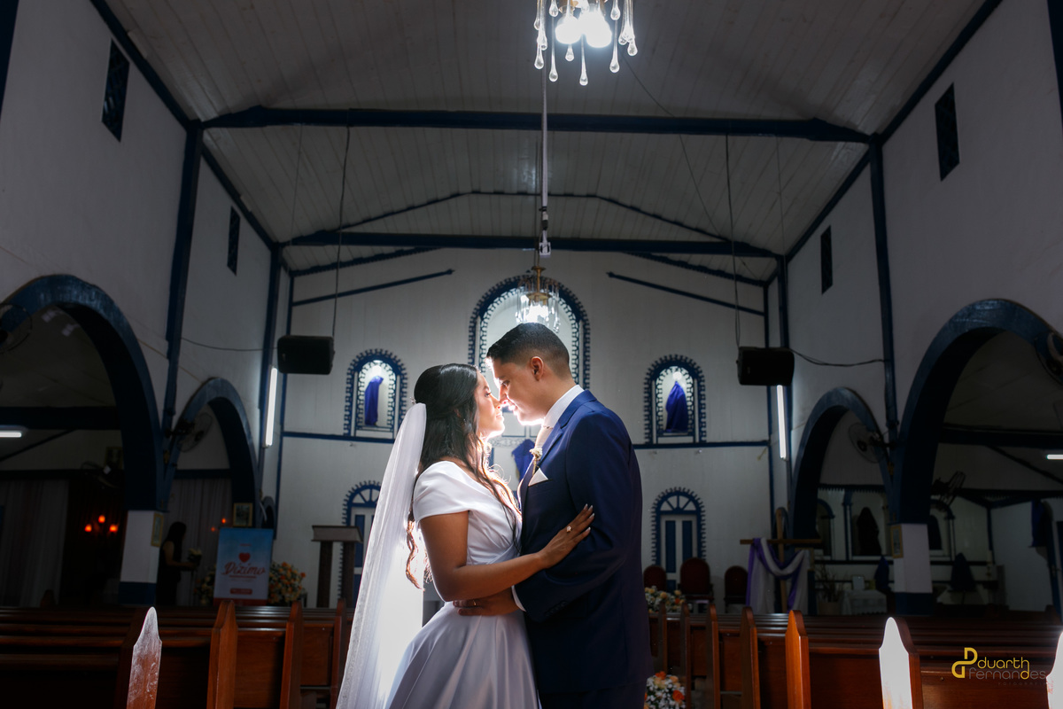 Noivos Gardênia e Arthur abraçados e sorrindo dentro da igreja iluminada após a cerimônia de casamento. Fotografia profissional por fotógrafo em Aricanduva.