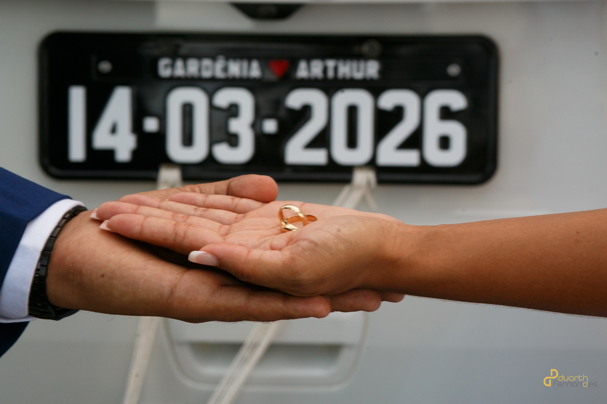 Detalhe das alianças de casamento de ouro sobre as mãos dadas dos noivos, com a placa decorativa do carro ao fundo marcando a data 14-03-2026. Casamento em Aricanduva.