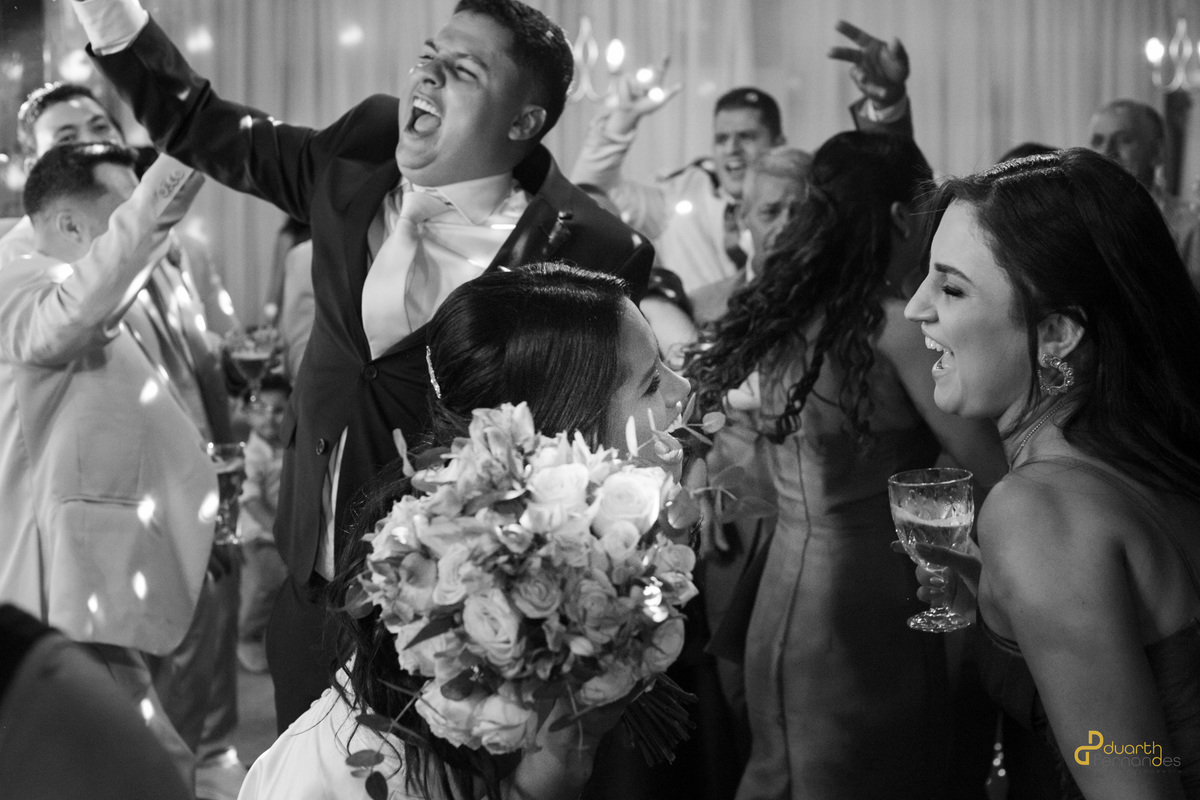 Foto em preto e branco capturando a noiva e os convidados rindo e cantando na pista de dança da recepção. Festa de casamento animada em Aricanduva, Minas Gerais.