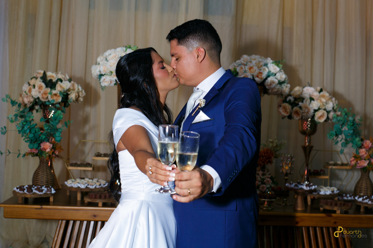 Noivos Gardênia e Arthur se beijando e brindando com taças de espumante em frente à mesa do bolo decorada com flores. Recepção de casamento em Aricanduva.