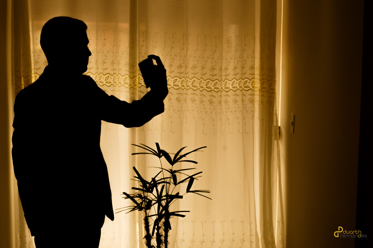 Silhueta do noivo Arthur segurando um frasco de perfume contra a cortina iluminada no making of. Fotografia criativa de casamento em Aricanduva.