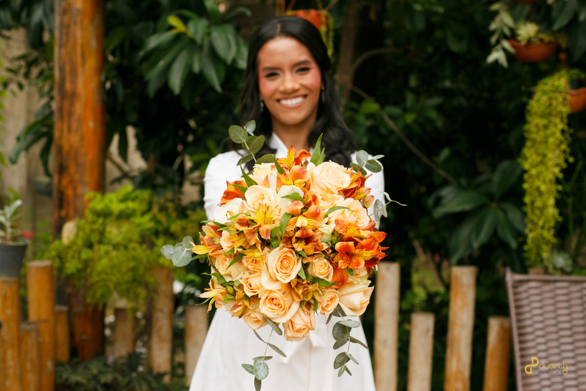 Noiva Gardênia sorridente em seu robe branco, segurando um lindo buquê de rosas e alstroemérias em tons de laranja e salmão no casamento em Aricanduva.