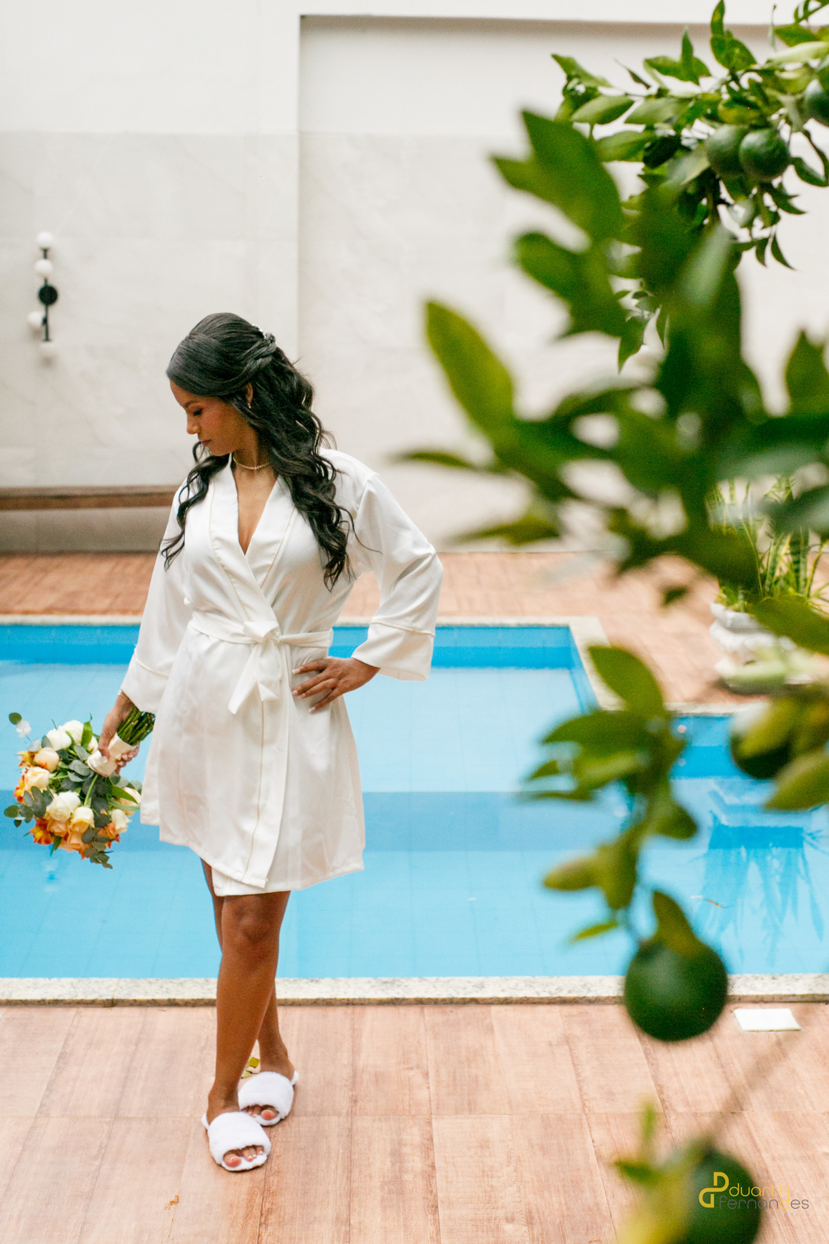 Noiva Gardênia de corpo inteiro à beira da piscina, usando robe e chinelos brancos enquanto segura o buquê de noiva. Preparativos para o casamento em Aricanduva, MG.