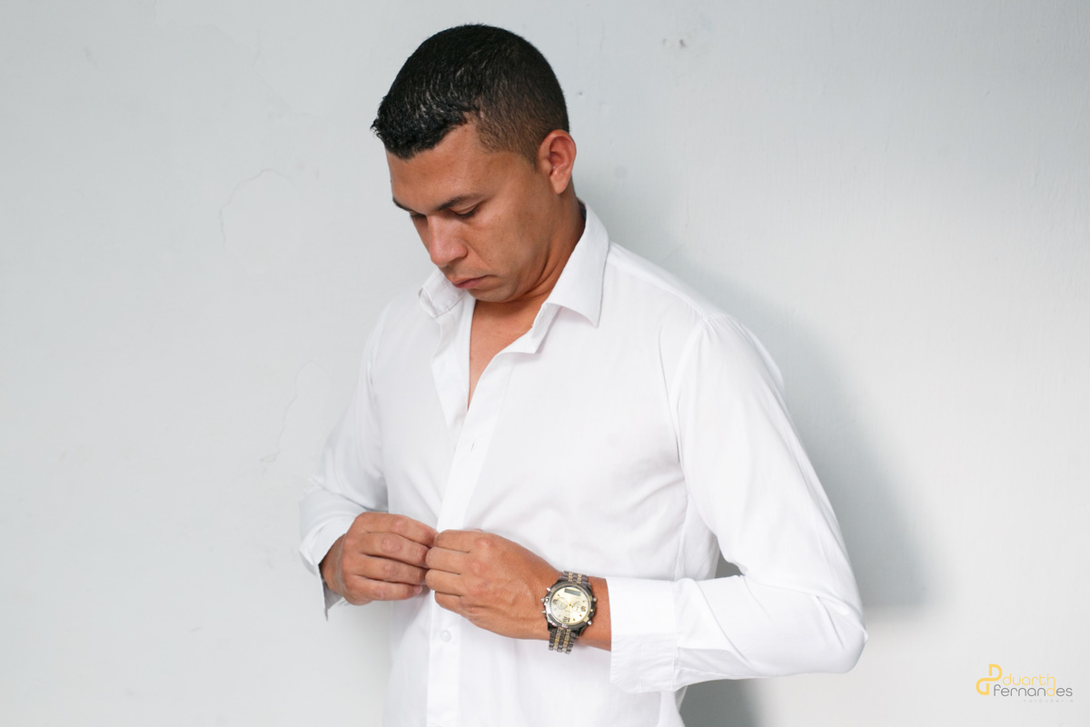 Noivo Sebastião em ensaio de making of abotoando sua camisa branca, destacando o relógio de pulso antes da cerimônia. Registro profissional realizado por fotógrafo de casamento em Angelândia, MG.