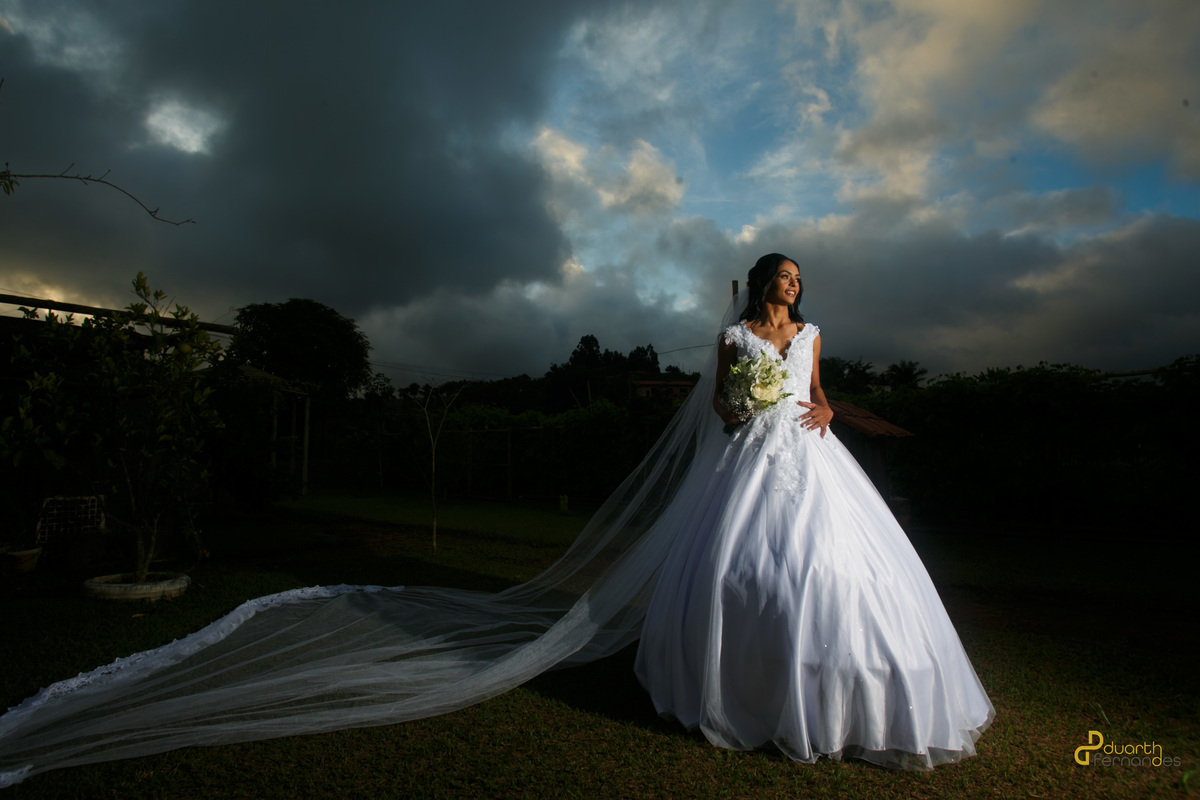Fotografia de casamento ao ar livre mostrando a noiva Jocilane de corpo inteiro com seu longo vestido branco rodado e véu estendido na grama sob um céu nublado ao entardecer em Angelândia.