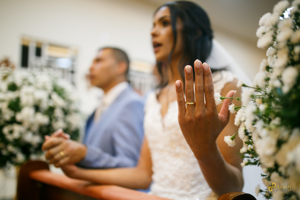 Noiva Jocilane com a mão levantada exibindo a aliança de casamento de ouro no altar da igreja, com o noivo Sebastião em segundo plano. Fotografia profissional de casamento em Angelândia.