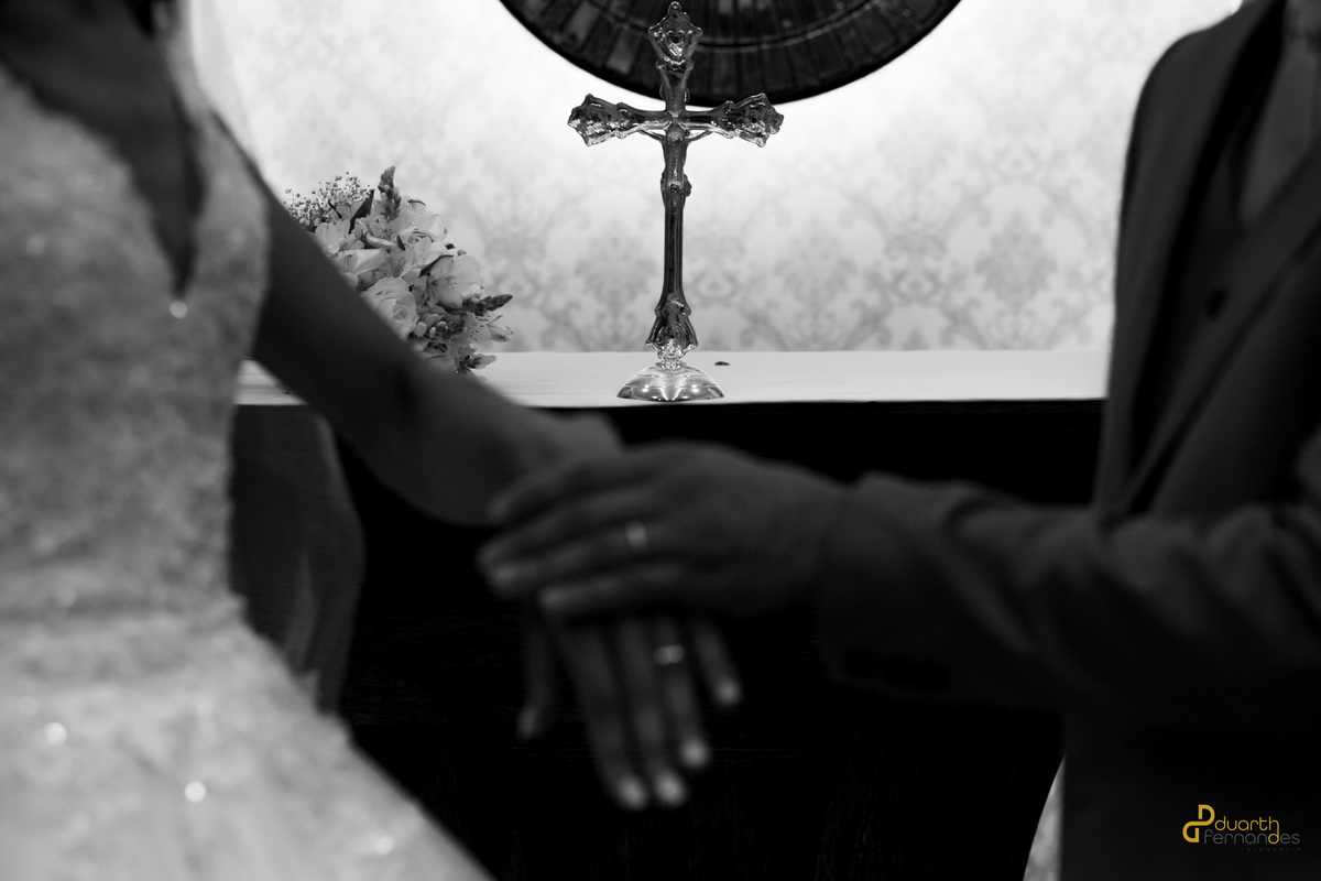 Noivo Sebastião abraçando a noiva Jocilane por trás e sorrindo no altar da igreja, ladeados por arranjos de flores brancas. Momento de alegria no casamento em Angelândia, MG.