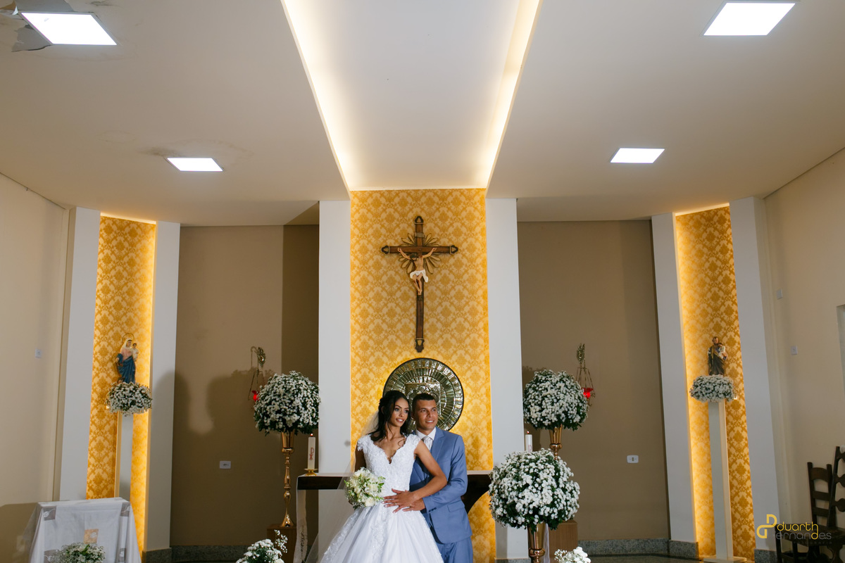 Detalhe em primeiro plano e preto e branco das mãos dadas dos noivos Jocilane e Sebastião no altar, com um crucifixo ao fundo. Fotografia artística de casamento em Angelândia.