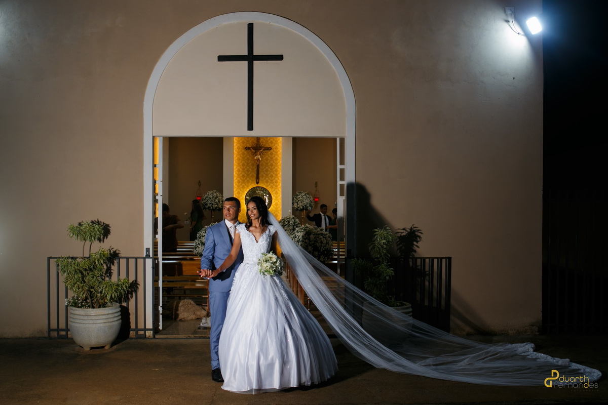 Noiva Jocilane com longo véu estendido posando de mãos dadas com o noivo Sebastião na entrada da igreja iluminada à noite. Ensaio pós-cerimônia de casamento em Angelândia, MG.