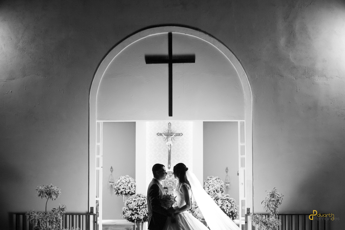 Silhueta em preto e branco dos noivos Sebastião e Jocilane se olhando na entrada da igreja abaixo de uma grande cruz. Fotografia conceitual de casamento em Angelândia.