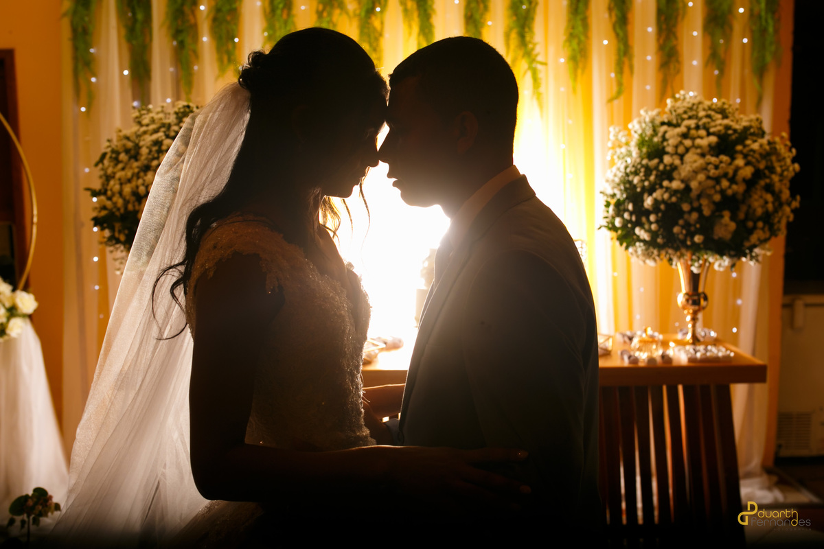 Retrato em contraluz destacando a silhueta da noiva Jocilane e do noivo Sebastião quase se tocando em frente à mesa do bolo iluminada. Ensaio de casamento em Angelândia.