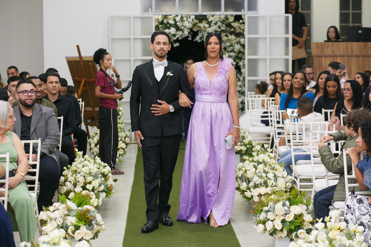 Entrada do noivo Dieique de terno preto caminhando sorridente pelo tapete do corredor central ao lado de sua mãe de vestido lilás. Início da cerimônia de casamento em Capelinha - MG.