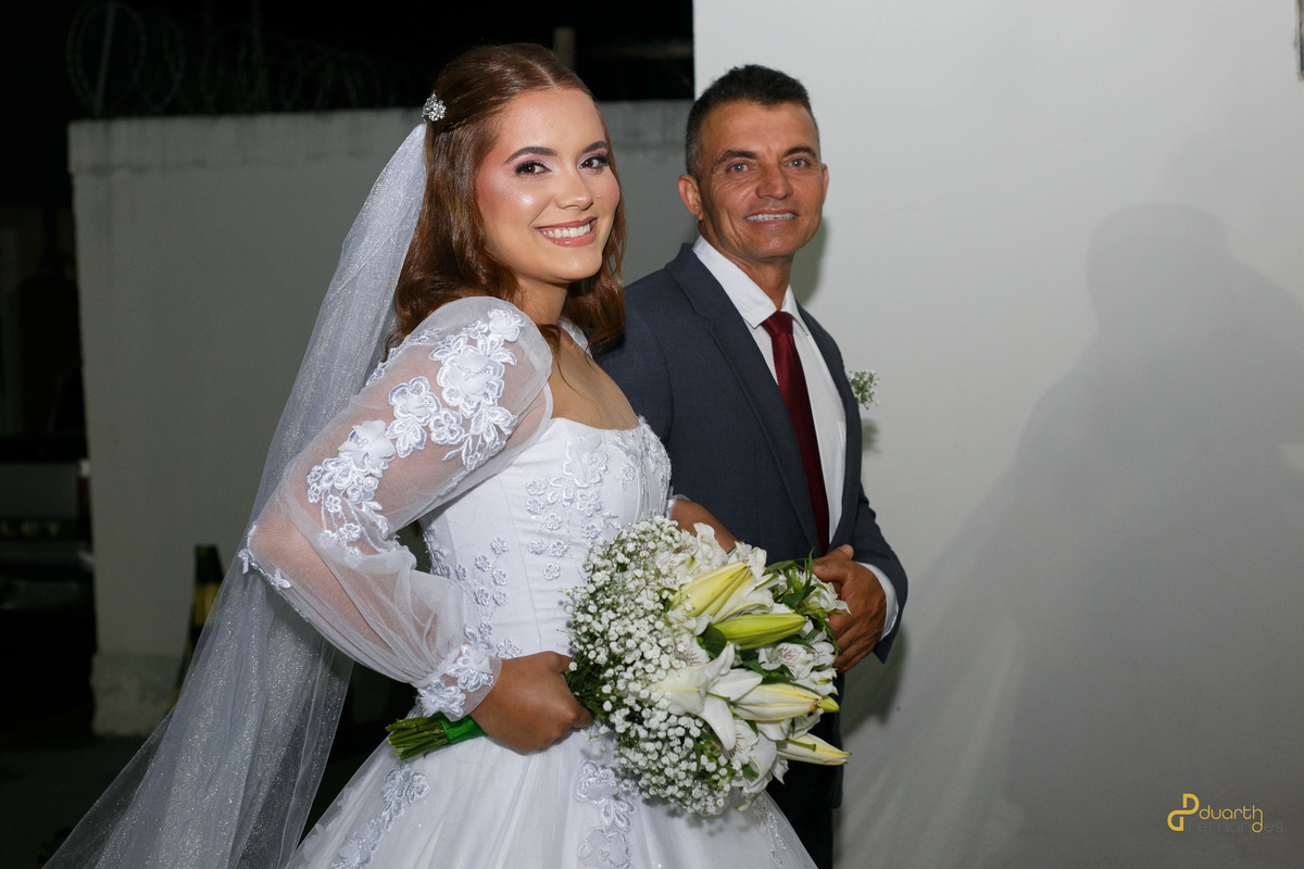 Plano médio aproximado da noiva Elen sorrindo no início do cortejo, caminhando de braços dados com seu pai para a cerimônia de casamento em Capelinha.