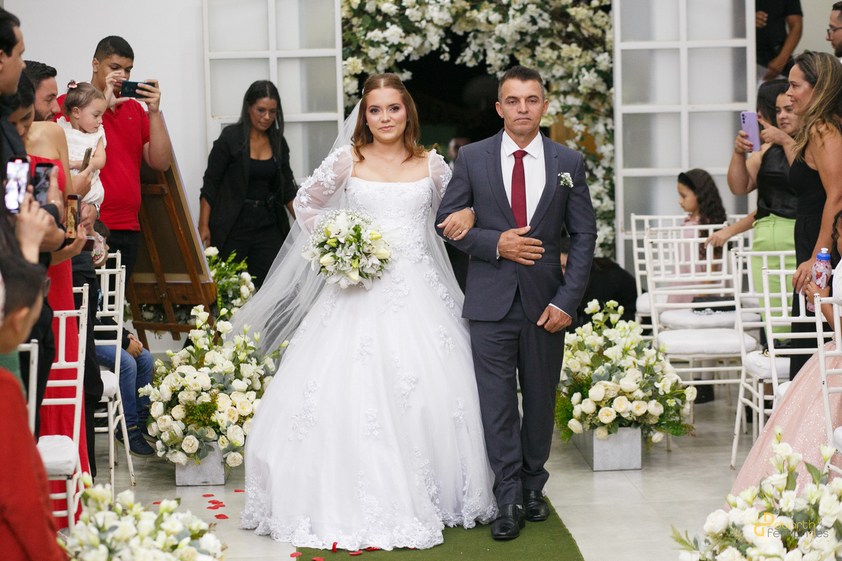 Entrada triunfal da noiva Elen de braços dados com seu pai na igreja, caminhando em direção ao altar ladeada por arranjos de flores brancas. Cerimônia de casamento em Capelinha - MG.