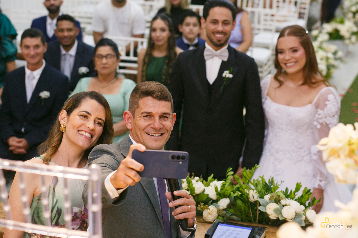 Casamento Capelinah Dieique Elen19_2.jpg: Padrinhos ou celebrantes sorrindo na frente do altar enquanto fazem uma foto selfie de celular, registrando os noivos Elen e Dieique ao fundo na igreja. Descontração na cerimônia em Capelinha - MG.