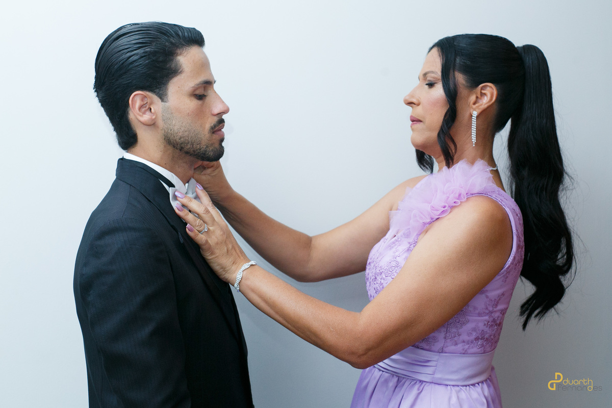 Noivo Dieique recebendo ajuda da mãe para ajustar a gravata borboleta no making of do noivo. Fotografia de casamento em Capelinha com foco na emoção dos preparativos familiares.