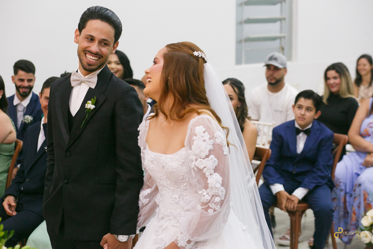 Noivos Elen e Dieique sorrindo e trocando olhares cúmplices de felicidade no altar. Registro espontâneo feito por fotógrafo de casamento em Capelinha - MG.