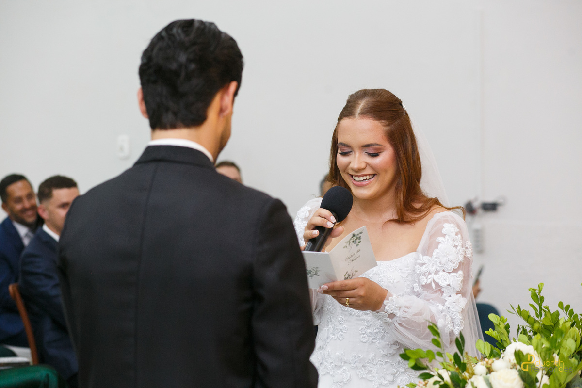 Noiva Elen sorridente e emocionada segurando o microfone e um papel com os votos de casamento na igreja. Detalhe do penteado e maquiagem de noiva em Capelinha - MG.