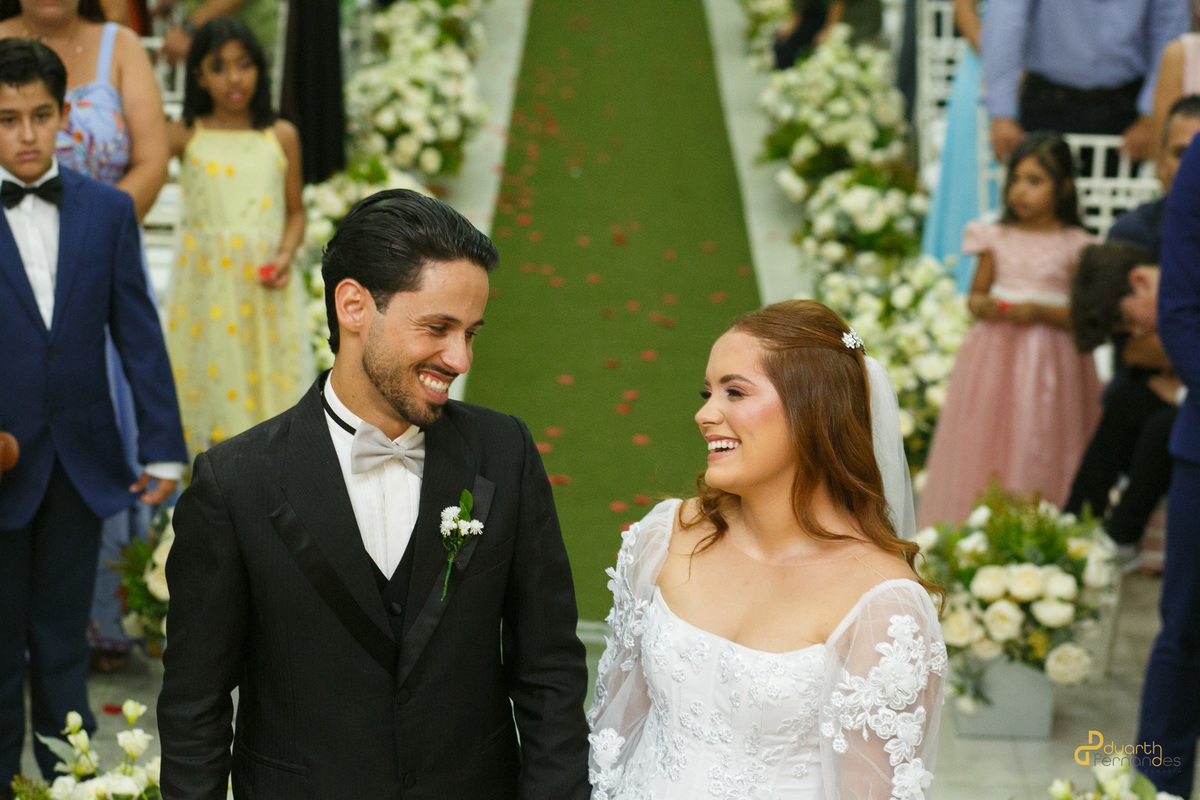 Noivos Dieique e Elen sorrindo um para o outro enquanto caminham no presbitério. O fundo desfocado mostra as crianças do cortejo na igreja de Capelinha - MG.