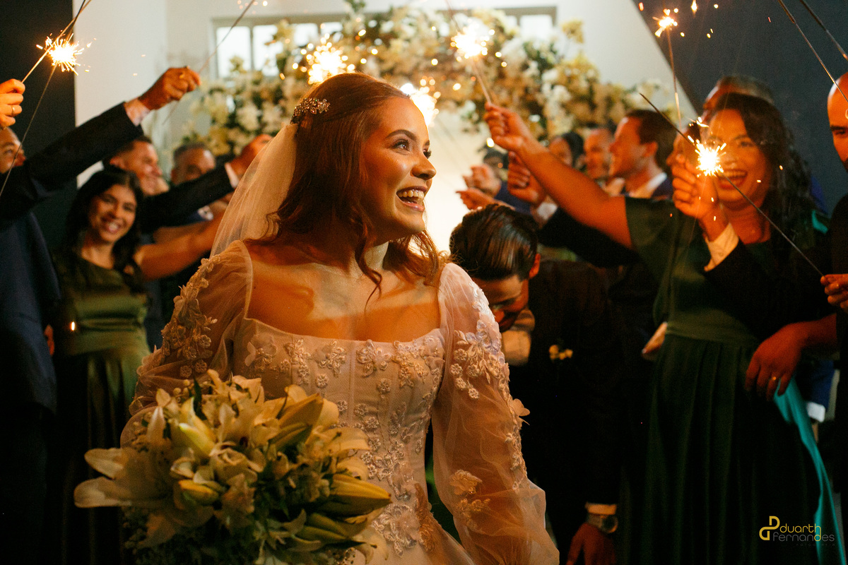 Noiva Elen sorridente caminhando no corredor da igreja ladeada por padrinhos segurando sparkles acesos. Detalhe da maquiagem da noiva iluminada e da cerimônia em Capelinha.