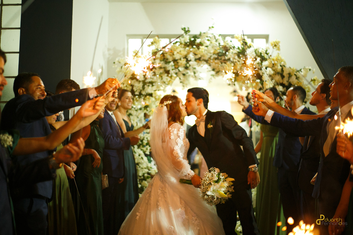 Noivos Dieique e Elen se beijando no corredor central da igreja sob o brilho de sparkles acesos pelos padrinhos. Registro emocionante de cerimônia de casamento em Capelinha - MG.