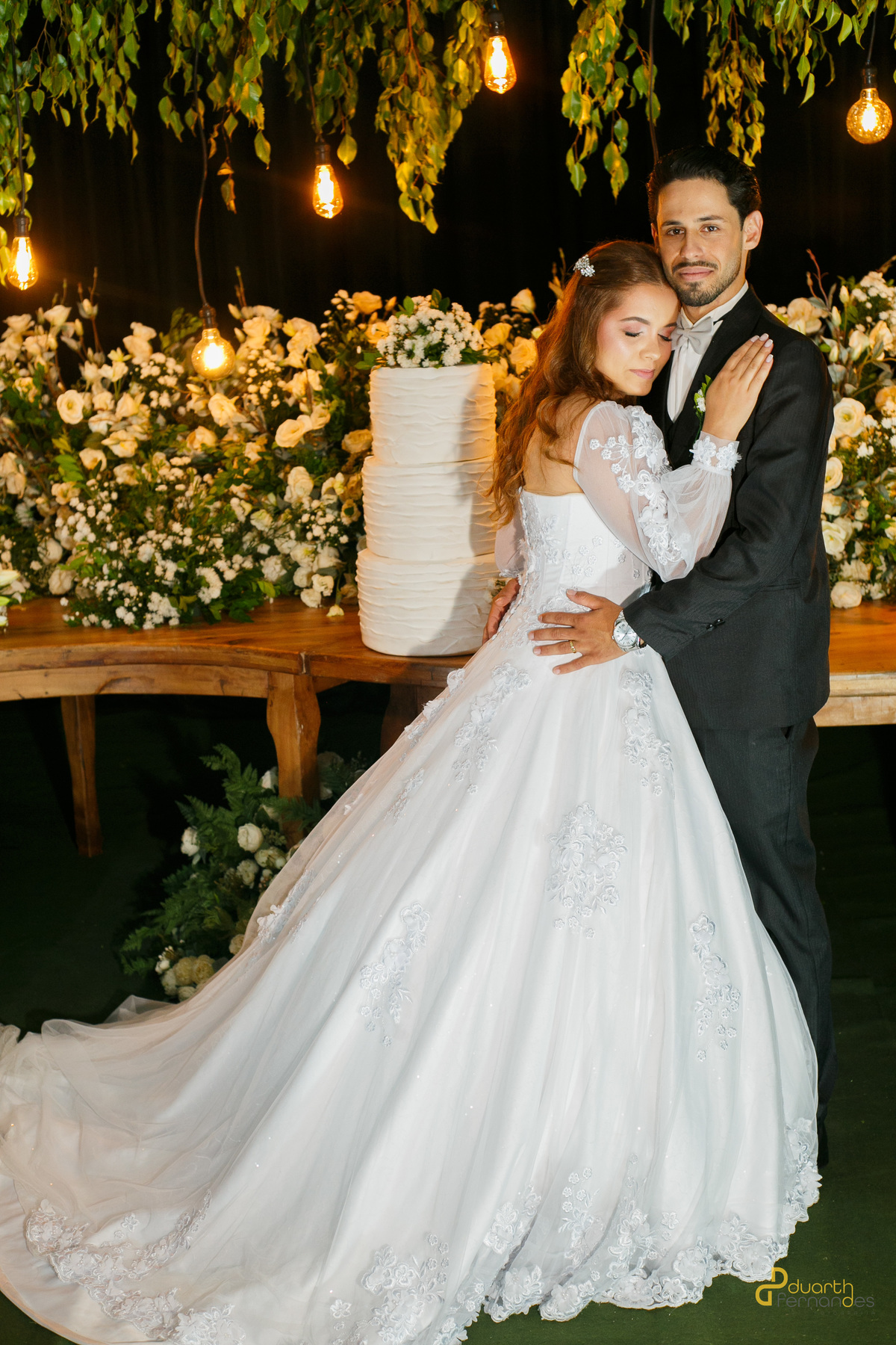 Noiva Elen e noivo Dieique abraçados em frente à mesa do bolo de casamento, decorada com arranjos de flores brancas e lâmpadas de filamento suspensas. Espaço de festas em Capelinha.