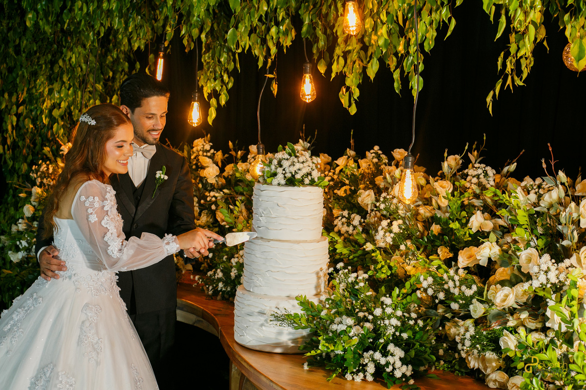 Noivos Dieique e Elen sorrindo juntos enquanto realizam o corte simbólico do bolo de casamento na recepção. Cobertura fotográfica completa de casamento em Capelinha - MG.