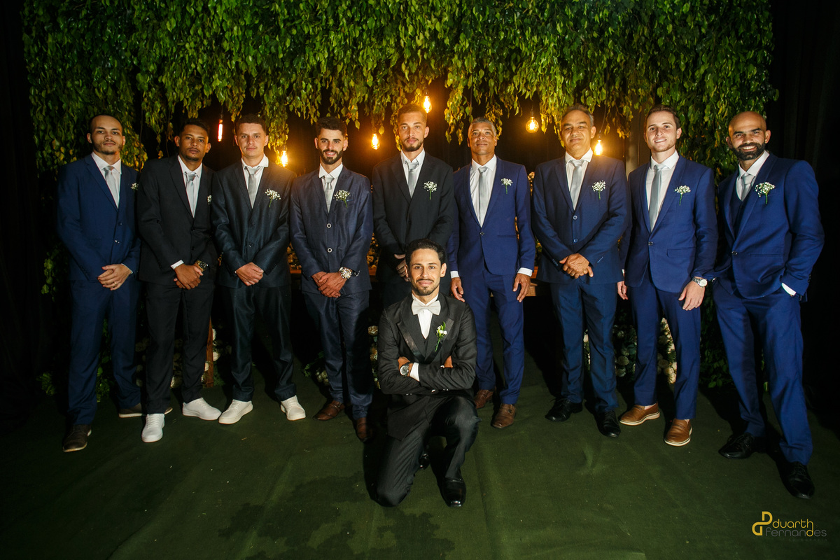 Noivo Dieique de braços cruzados e ajoelhado à frente de sua bancada de padrinhos, todos vestindo ternos azuis alinhados na recepção do casamento em Capelinha - MG.