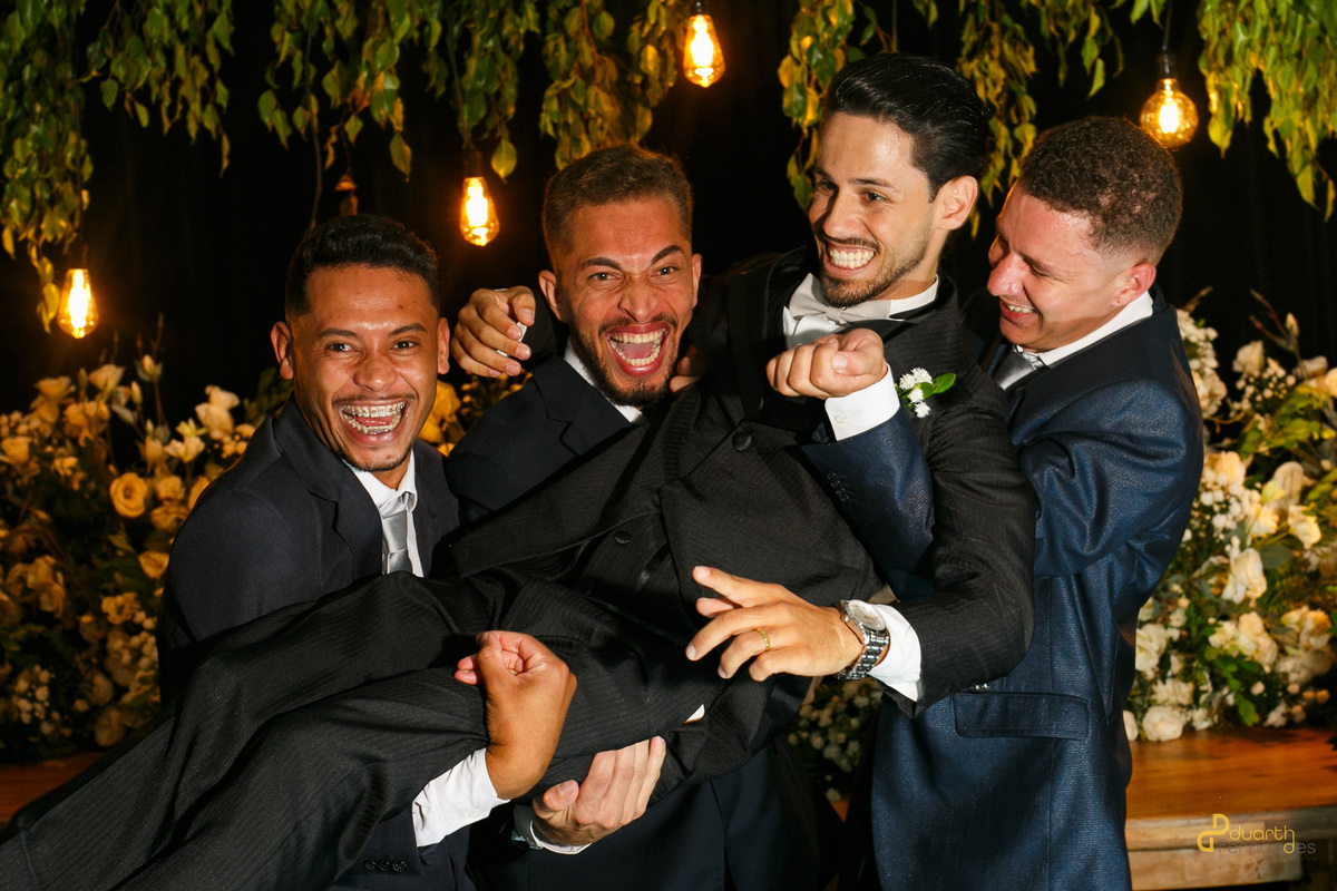Registro descontraído do noivo Dieique sendo carregado no colo e jogado para o alto por seus padrinhos durante a festa de recepção de casamento em Capelinha.