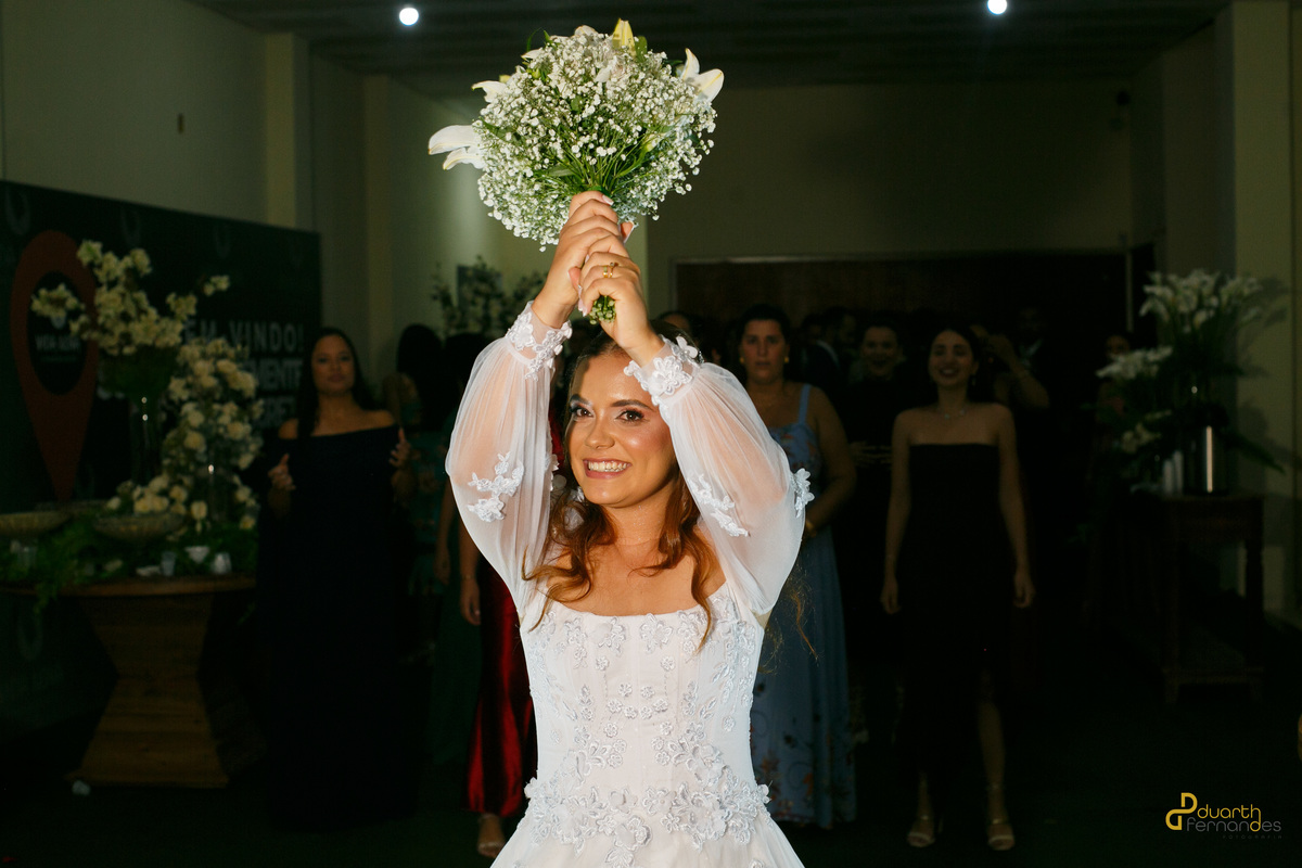 Noiva Elen de frente sorrindo com os braços erguidos, segurando o buquê de lírios brancos instantes antes de jogá-lo para as convidadas na festa em Capelinha - MG.