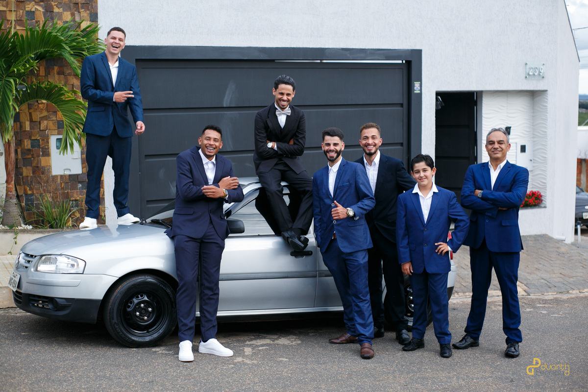 Foto criativa do noivo Dieique e seus padrinhos posando com ternos azuis e pretos ao redor de um carro prata. Ensaio divertido antes da cerimônia de casamento em Capelinha.