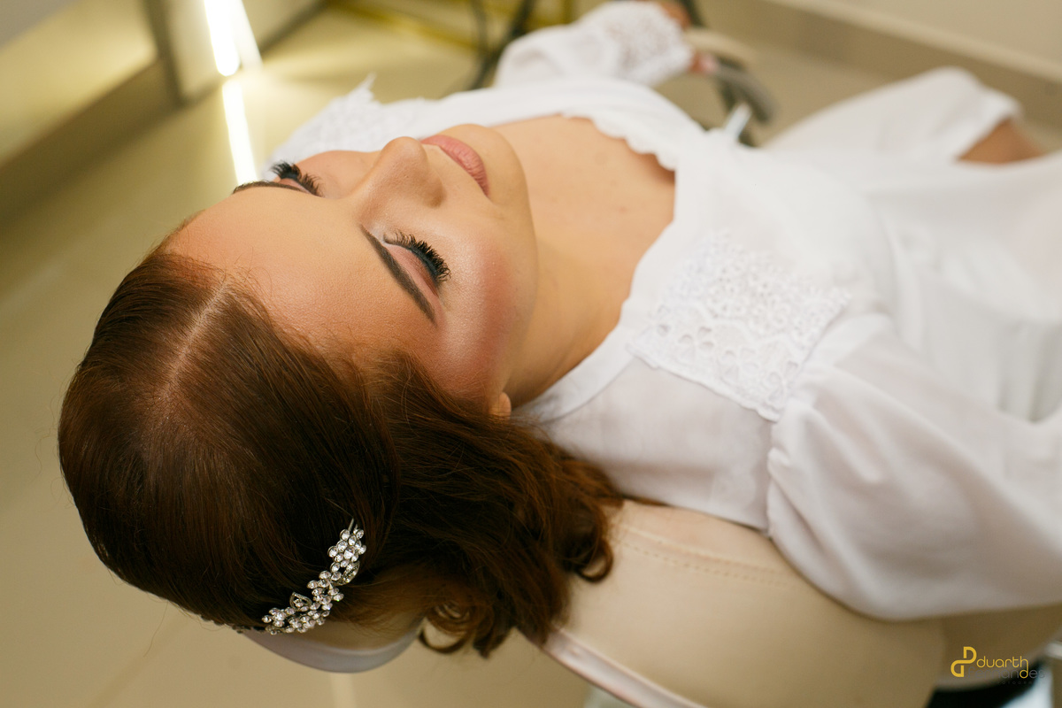 Noiva Elen de olhos fechados na cadeira do salão, exibindo o penteado com arranjo de brilhantes e a maquiagem da noiva finalizada. Dia de noiva e maquiagem em Capelinha - MG.