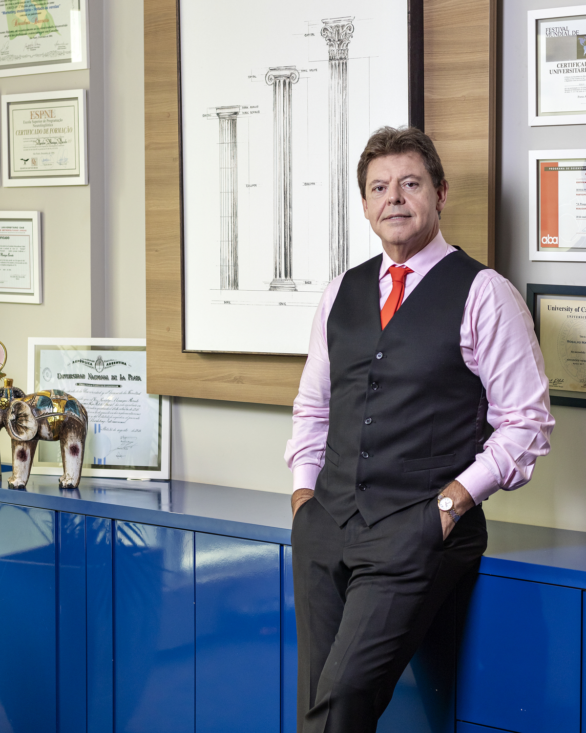 Em pé, apoiado em estante, de colete preto.