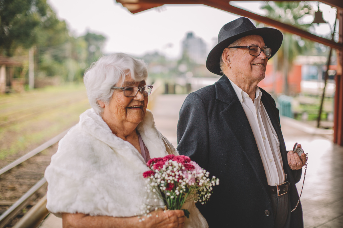 Idosa de cabelos brancos com um buquê na mão de mãos dadas com um senhor idoso que está de chapéu preto e segurando um relógio antigo