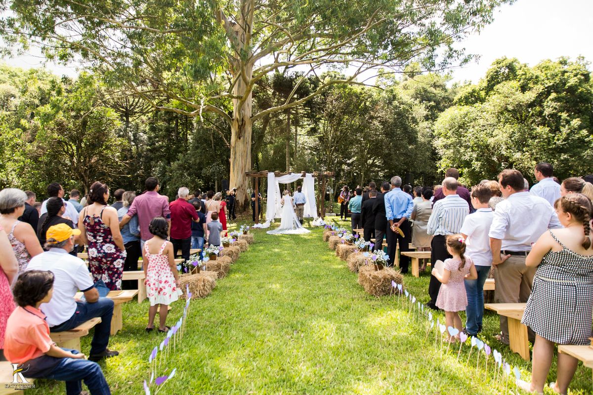 casamento ao livre em francisco beltrão cabanha manhare de thairine e luciano katy tesser fotografa os noivos entrando na cerimonia religiosa vendo a decoração do casamento ela de vestido de noiva e buquet e o noivo com traje gaucho