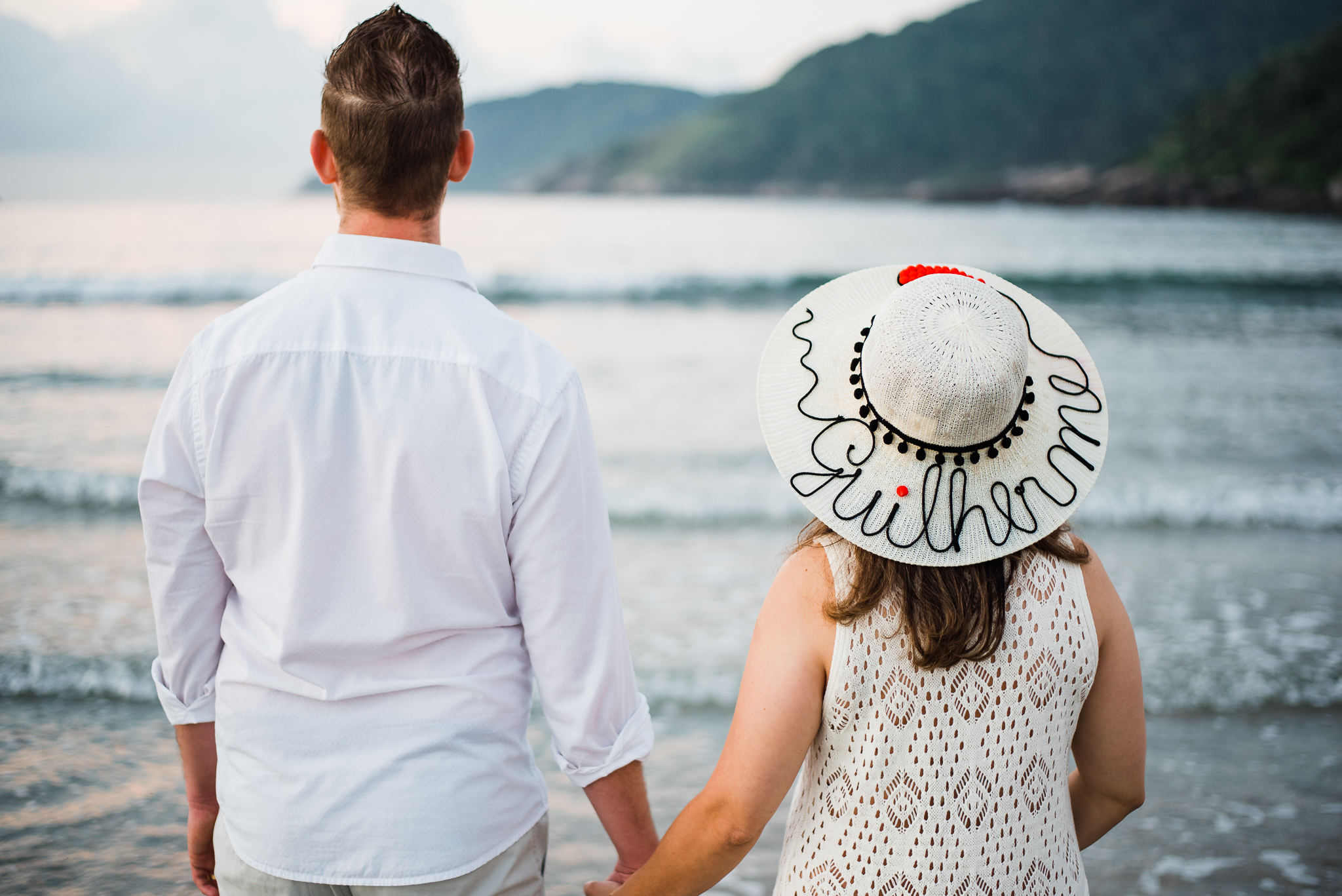 Ensaio gestante praia do Guarujá casal feliz abraçados litoral mar areia montanhas água salgada sol chapéu com o nome chapéu escrito  gestante parto maternidade gestação #gestante mamadeira amamentação barriga de gestante ensaio fotográfico book