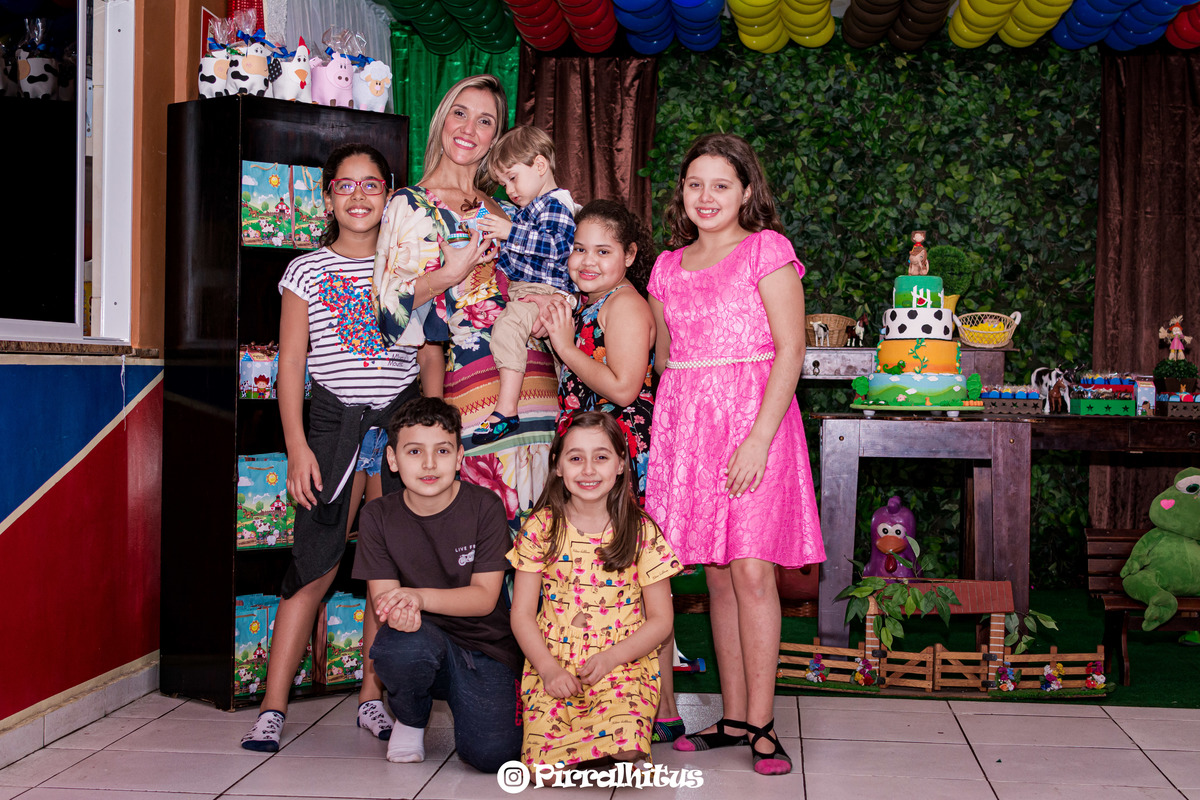 Gabriel no colo da mãe acompanhado de crianças em frente a mesa do bolo. 