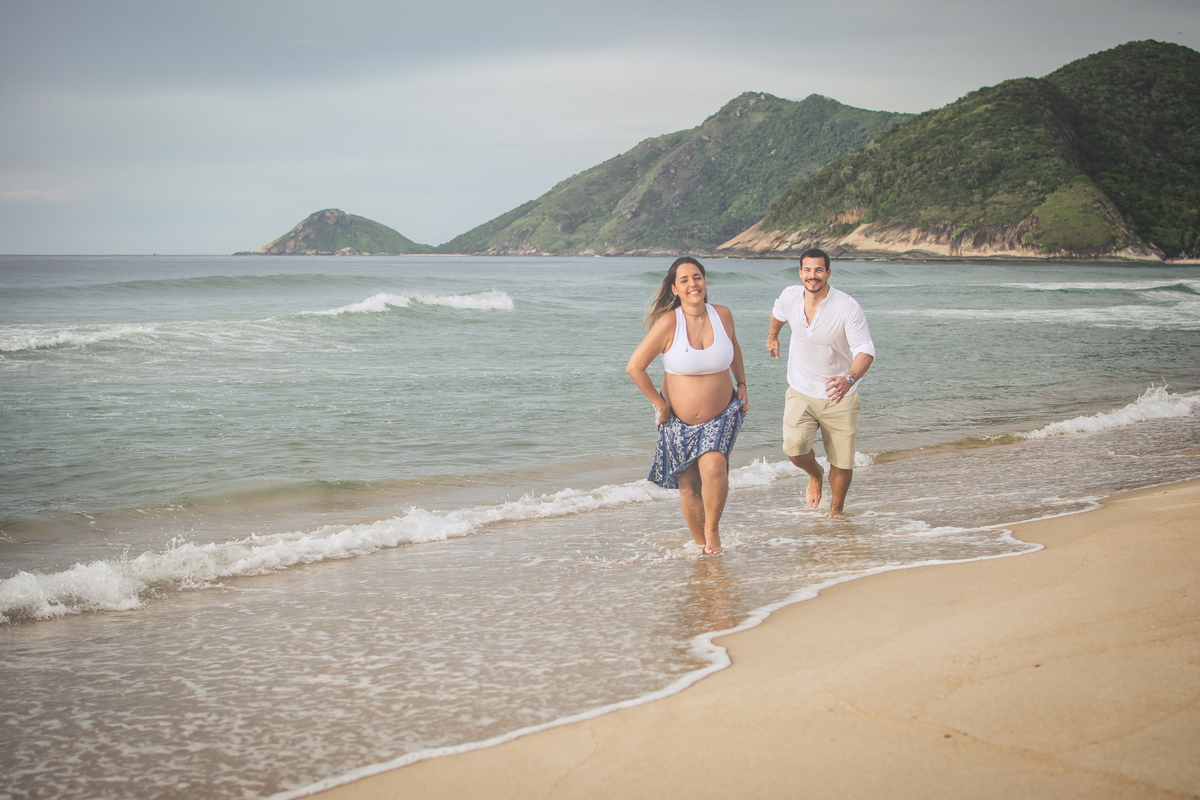 Casal correndo na areia da praia durante ensaio gestante romantico.