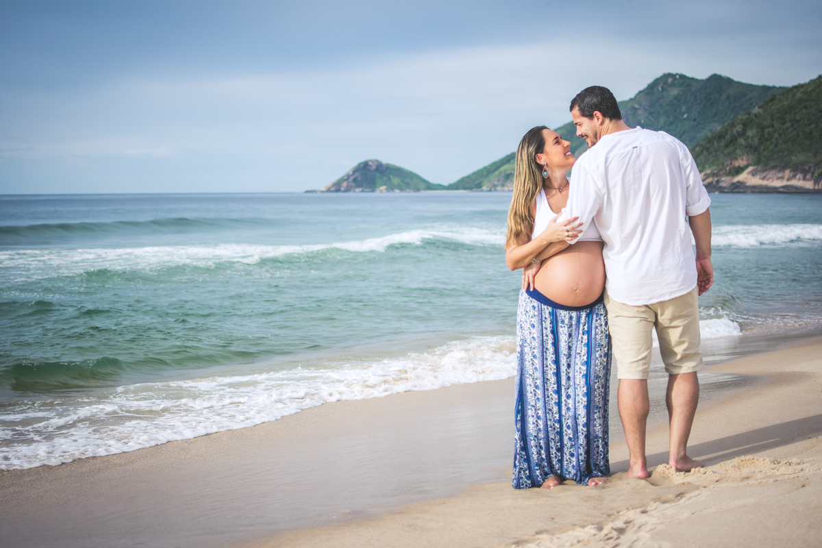 casal abraçado fazendo pose para ensaio fotográfico de gestante em grumari, ela veste saia estampada azul e croped branco, ele usa bermuda de sarja off-white e camisa de manga comprida branca.
