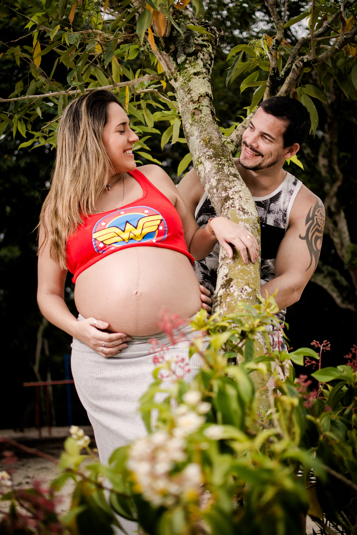 Casal brincando de se esconder atrás da árvore durante ensaio gestante no bosque da barra, ela usa saia bege e camiseta da mulher maravilha e ele usa camisa regata preta e bermuda jeans.