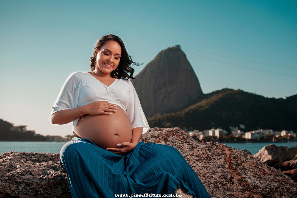Gestante no aterro do Flamengo sentada na pedra, em frente ao Pão de açúcar. Vestindo croped branco e saia azul tifany.