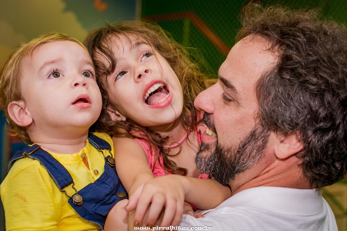 Pai carregando casal de filhos no colo no salão de festas infantis.