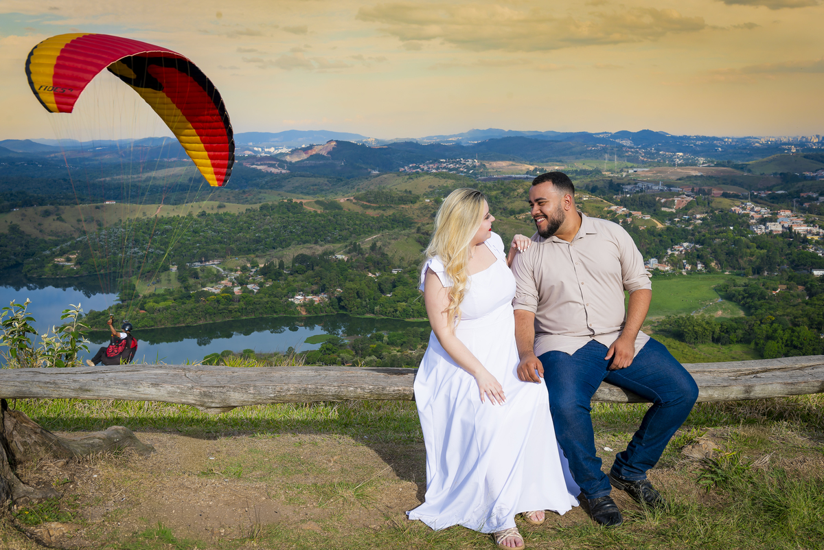 No alto do Morro do Capuava, Kamila e Daniel contemplam o amor que os eleva — tão leve quanto o voo ao fundo., ensaio pré-wedding Alphaville, ensaio fotográfico pré-wedding Morro do Capuava ensaio, fotos de casal ao pôr do sol, pré-wedding com parapente