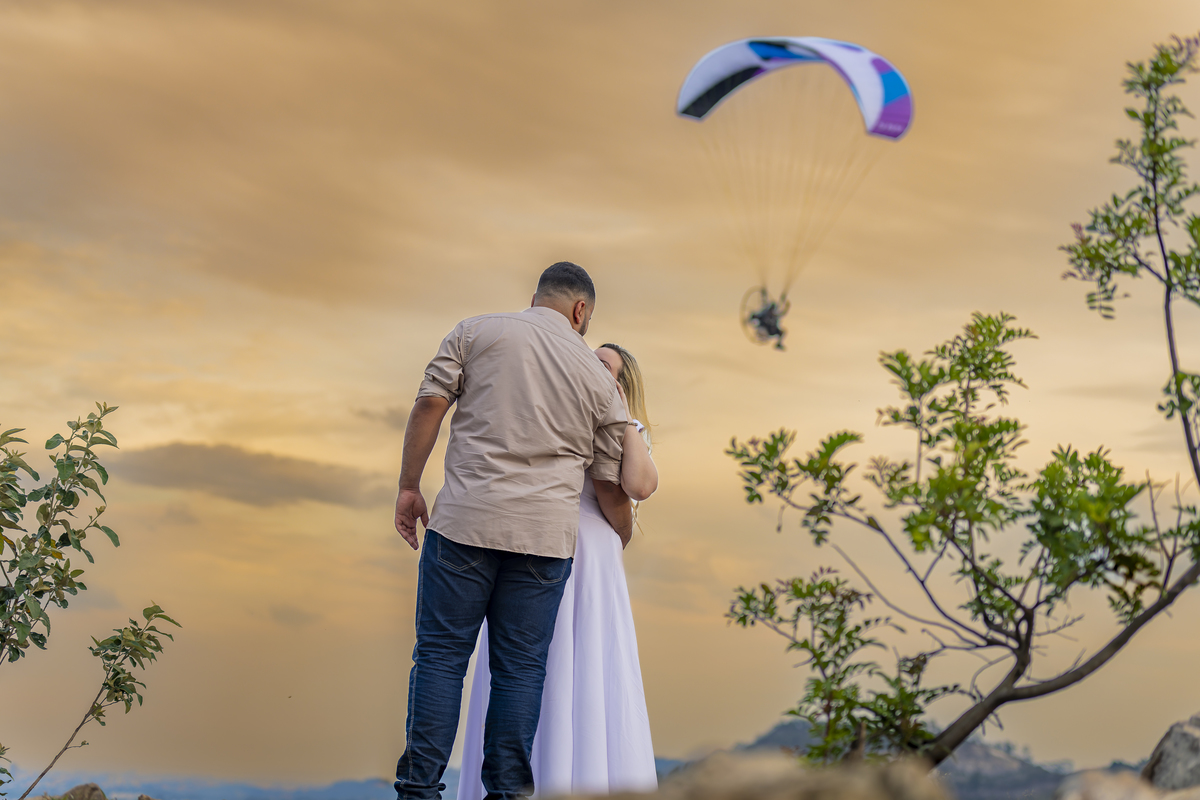 No alto do Morro do Capuava, Kamila e Daniel contemplam o amor que os eleva — tão leve quanto o voo ao fundo., ensaio pré-wedding Alphaville, ensaio fotográfico pré-wedding Morro do Capuava ensaio, fotos de casal ao pôr do sol, pré-wedding com parapente