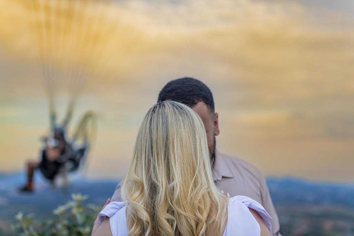 No alto do Morro do Capuava, Kamila e Daniel contemplam o amor que os eleva — tão leve quanto o voo ao fundo., ensaio pré-wedding Alphaville, ensaio fotográfico pré-wedding Morro do Capuava ensaio, fotos de casal ao pôr do sol, pré-wedding com parapente