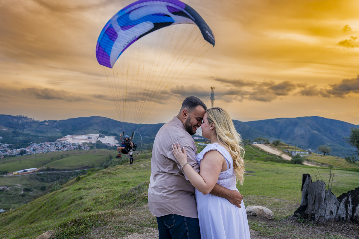 No alto do Morro do Capuava, Kamila e Daniel contemplam o amor que os eleva — tão leve quanto o voo ao fundo., ensaio pré-wedding Alphaville, ensaio fotográfico pré-wedding Morro do Capuava ensaio, fotos de casal ao pôr do sol, pré-wedding com parapente