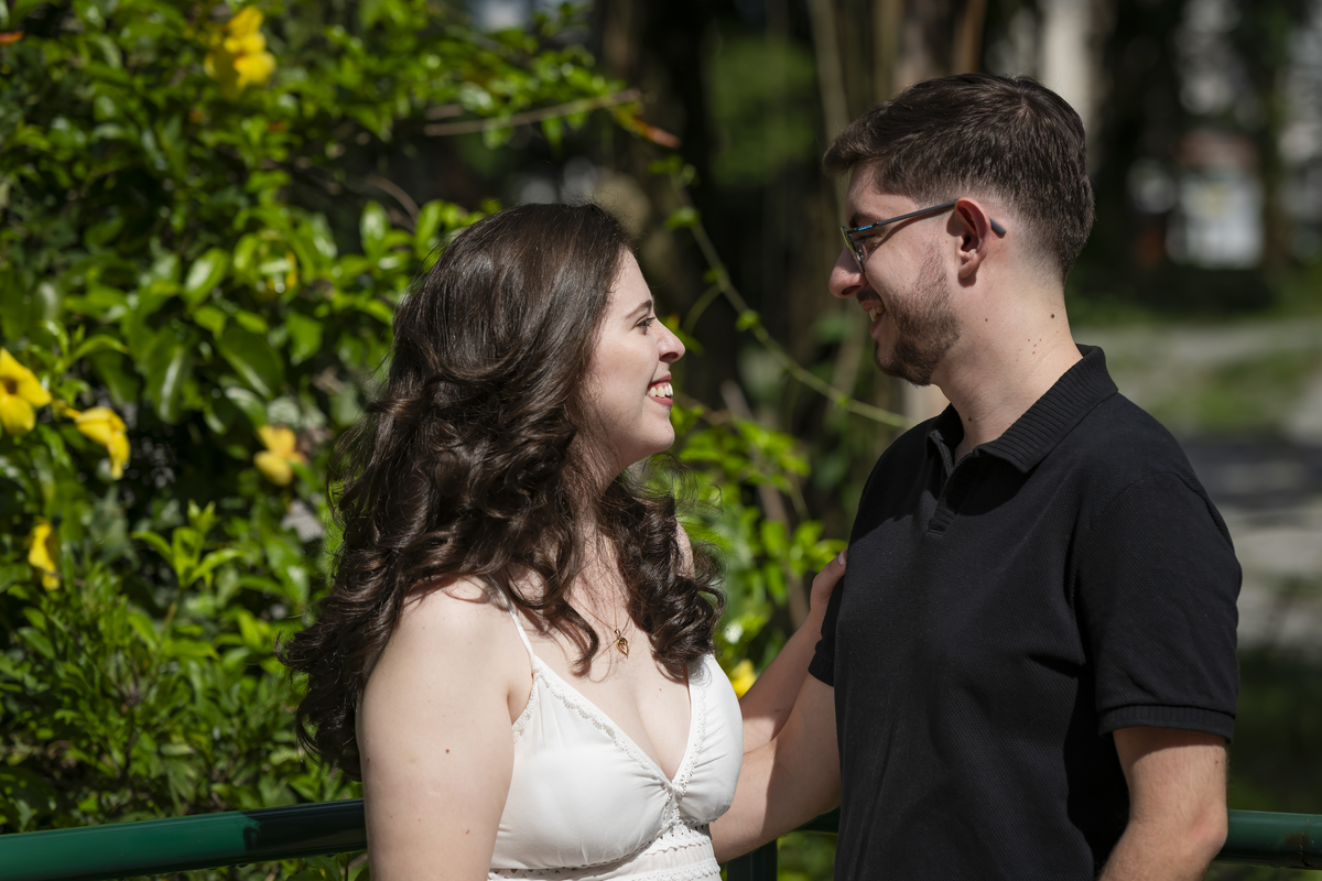 Casal sorridentes, olhar próximo e cúmplice, junto a um corrimão verde cercado por flores amarelas no Jardim Botânico de São Paulo.