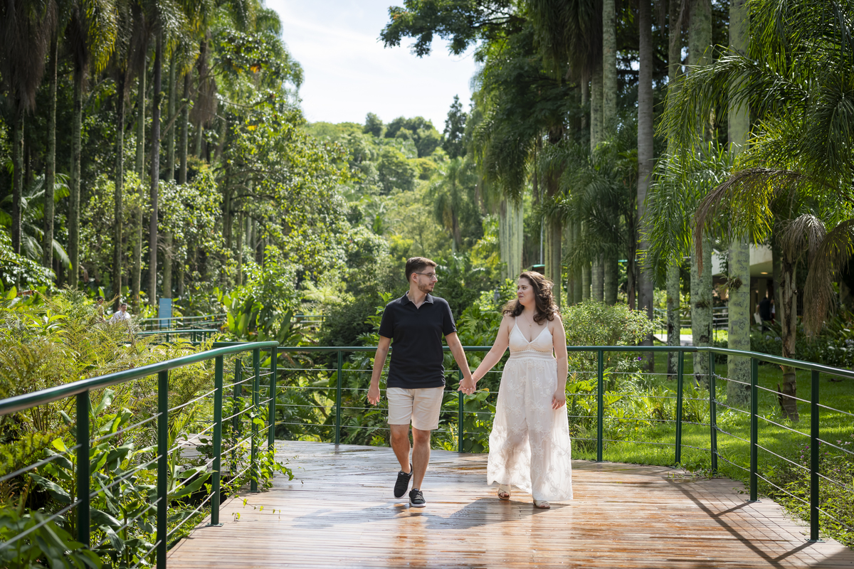 Casal caminham de mãos dadas por passarela de madeira em meio à vegetação tropical do Jardim Botânico de São Paulo.